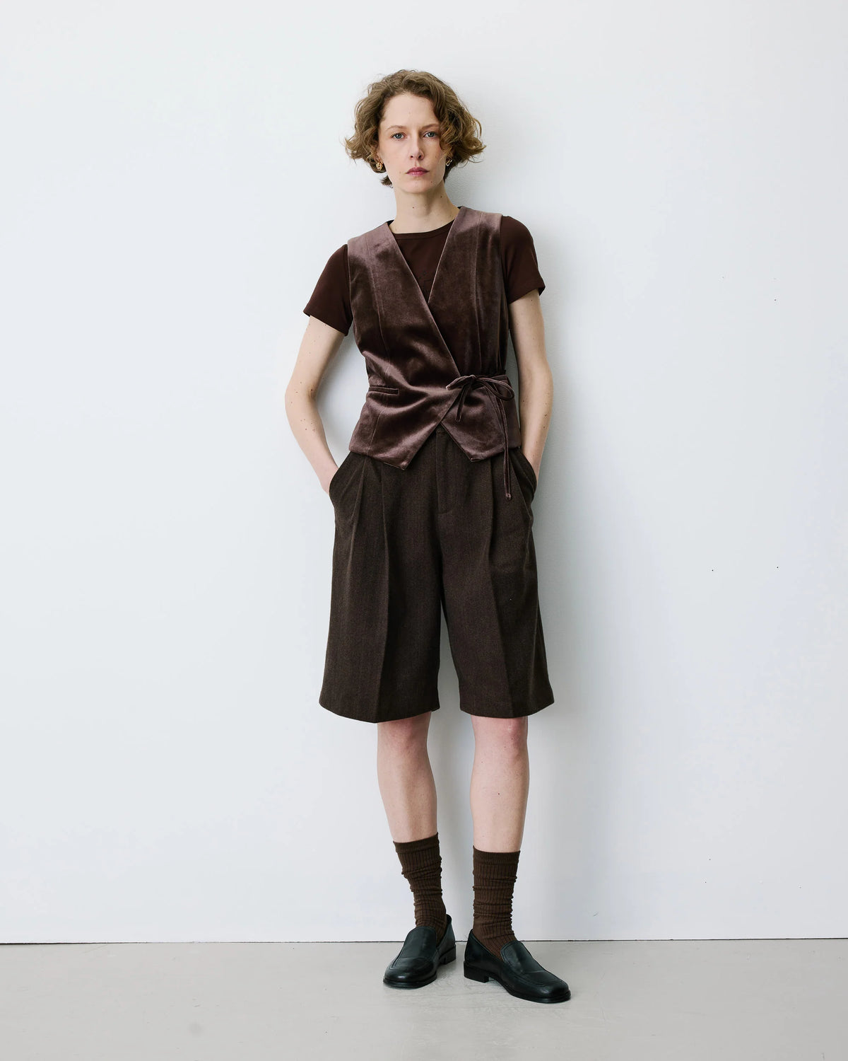 A model stands against a plain white wall wearing a dark brown velvet short-sleeve wrap top with a tied waist, matching dark brown pleated shorts, brown socks, and black shoes.
