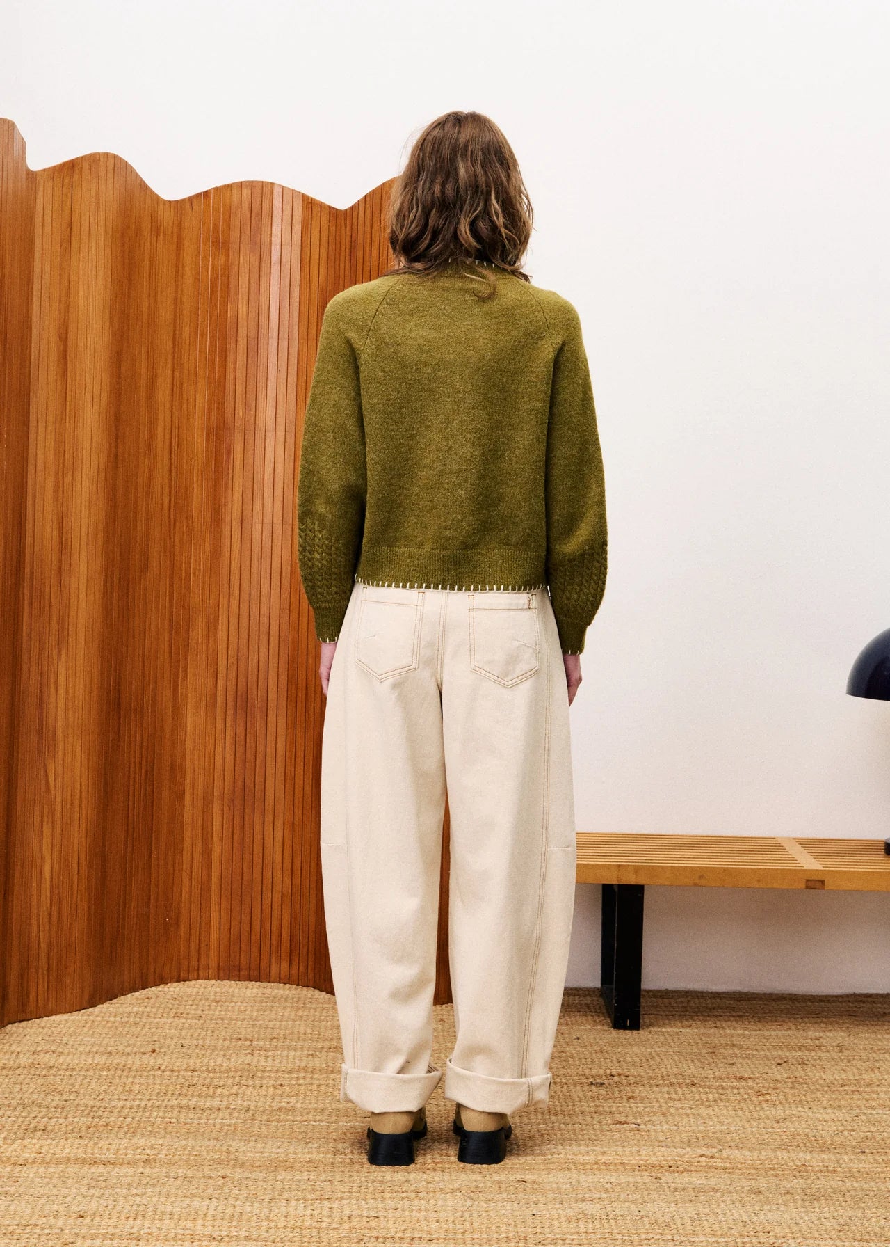 Person wearing a green sweater and white pants standing in front of a wooden screen.