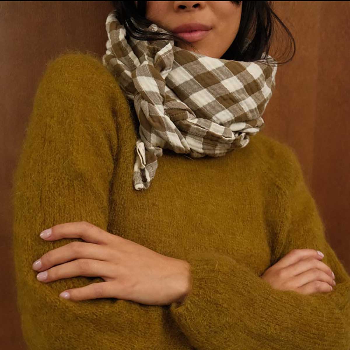 Person wearing a mustard yellow sweater and a plaid scarf against a brown background