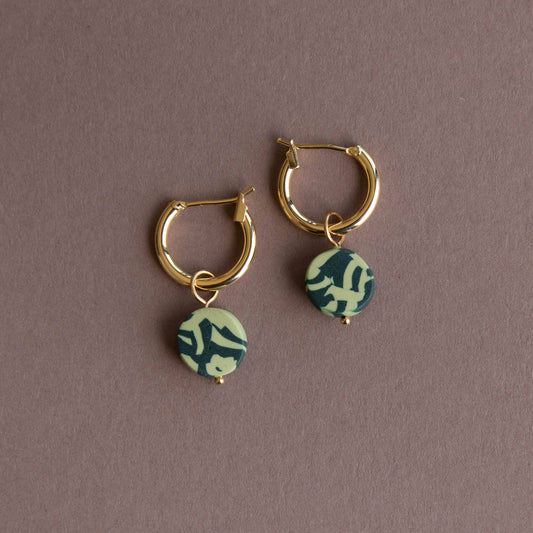 Gold hoop earrings with green and white patterned charms on a brown background