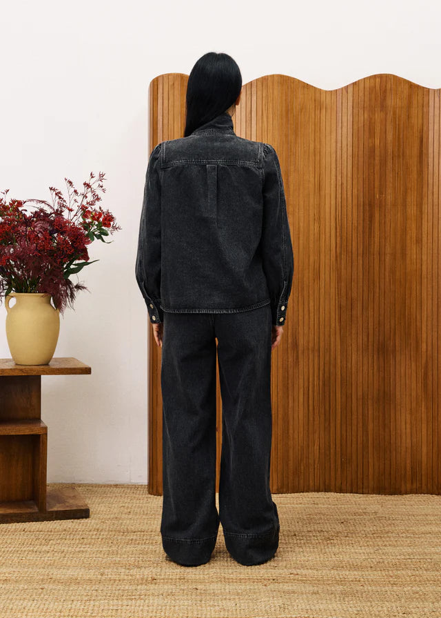Person wearing a black denim jacket and pants standing in front of a wooden screen with a vase of flowers on a table.