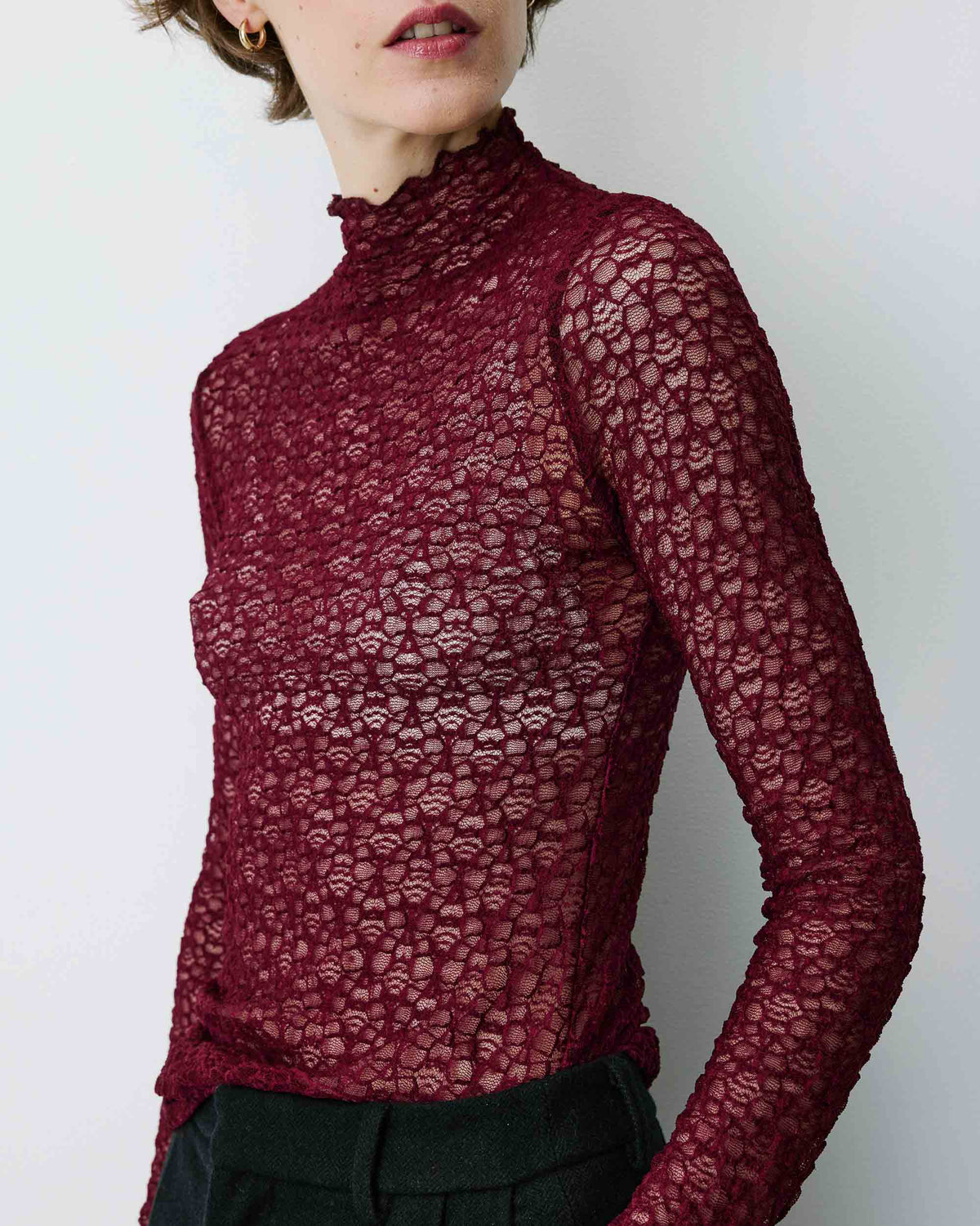 A woman wearing a long-sleeve, high-neck, sheer lace top in deep burgundy, paired with dark trousers, against a plain light gray background.