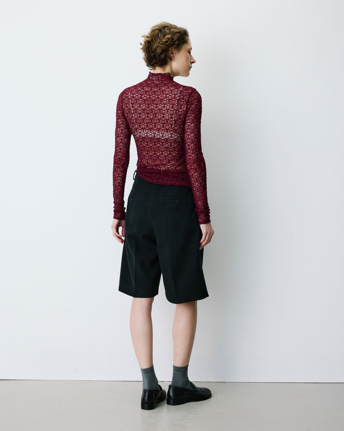A model shown from the back wearing a dark red patterned mock-neck long-sleeve top, black shorts, gray socks, and black shoes, standing against a plain white wall.