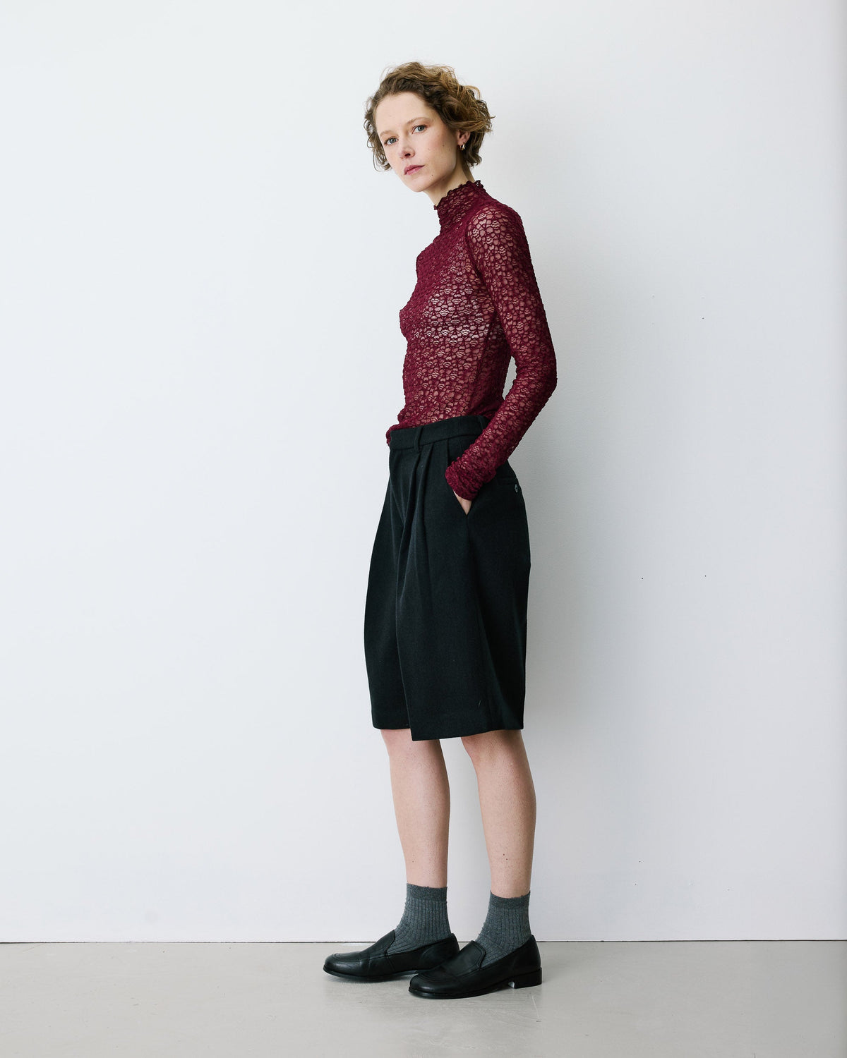 A model stands in profile wearing a burgundy long-sleeve lace mock-neck top, black pleated knee-length shorts, dark gray socks, and black leather loafers against a plain white studio wall.
