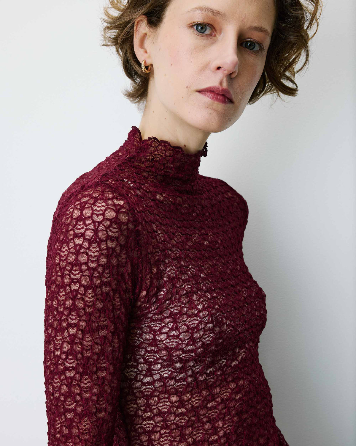 A woman wearing a burgundy lace mock-neck long-sleeve top, photographed against a plain light gray background.