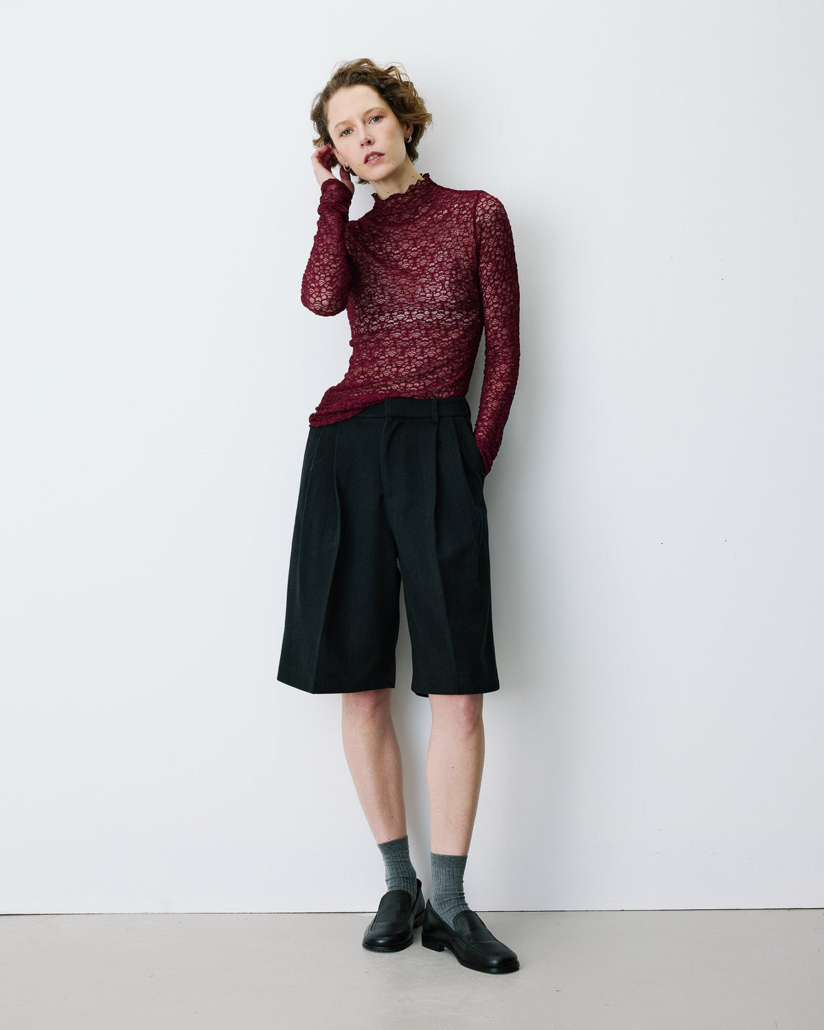 A model stands against a plain white wall wearing a burgundy textured mock-neck long-sleeve top, black pleated knee-length shorts, gray socks, and black leather shoes.