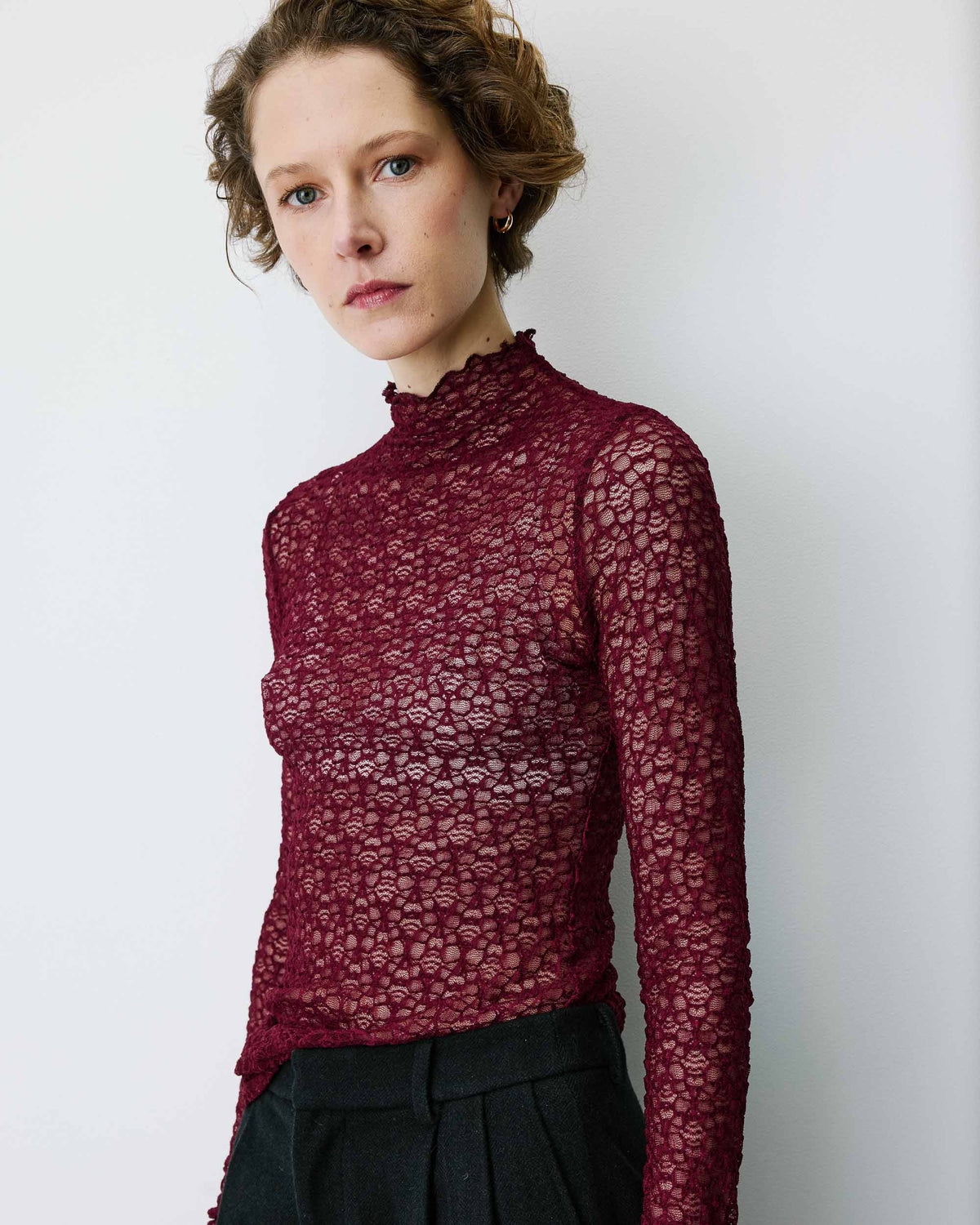 A woman wearing a sheer, long-sleeve lace mock-neck top in deep burgundy, paired with dark trousers, standing against a plain light gray wall.