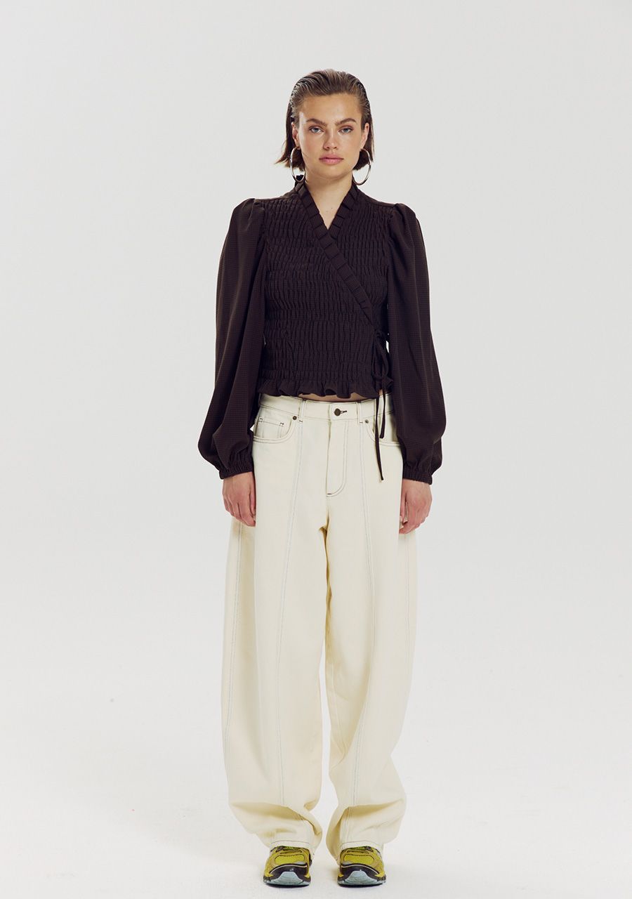A model wearing a A dark brown textured long-sleeve blouse with a mock neck, puff sleeves, and a drawstring waist, wearing cream trousers and trainers shown from the back on a light gray background.