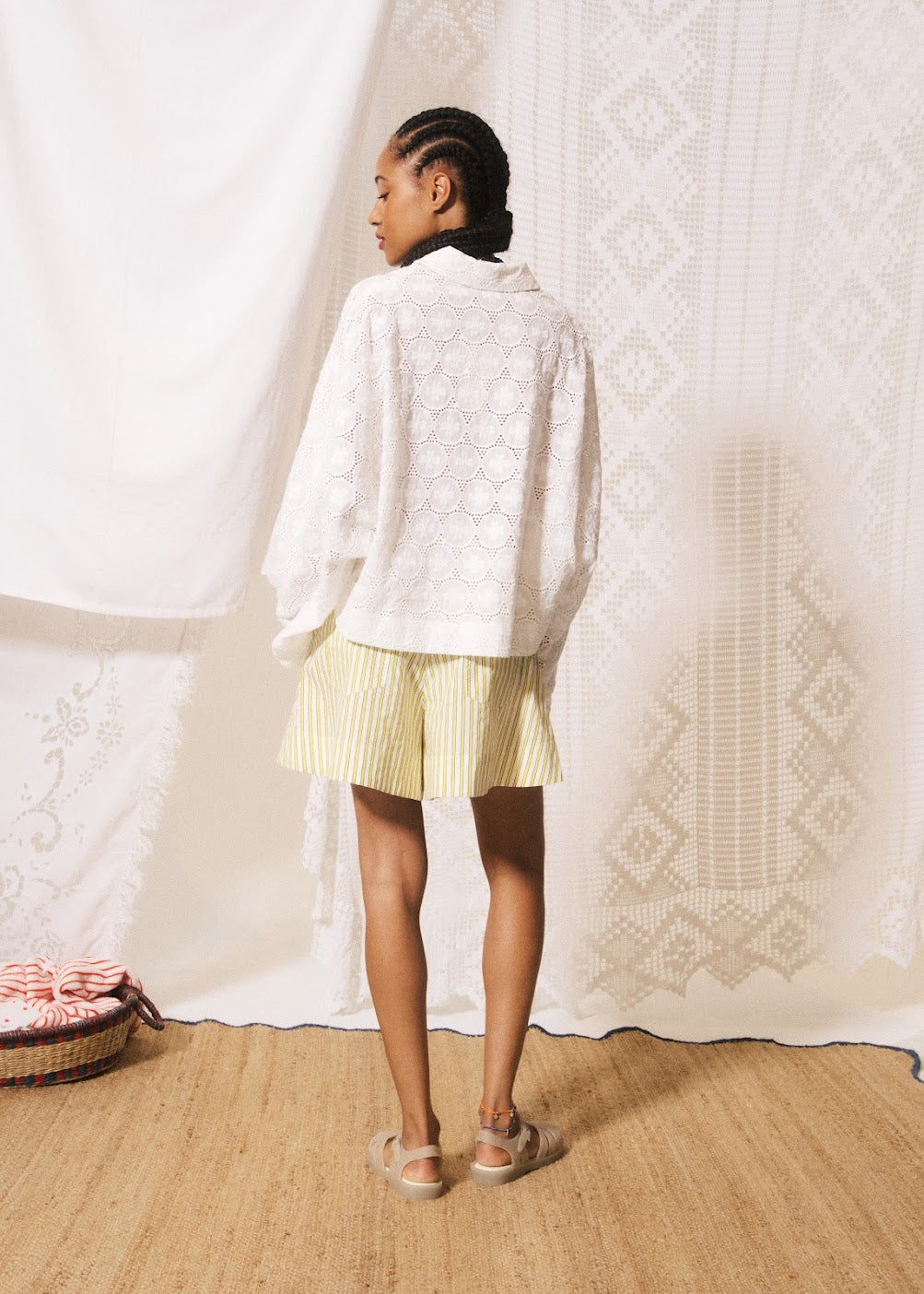 Person wearing a white cardigan and yellow shorts standing in front of a textured white curtain.