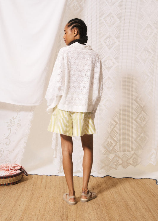 Person wearing a white cardigan and yellow shorts standing in front of a textured white curtain.