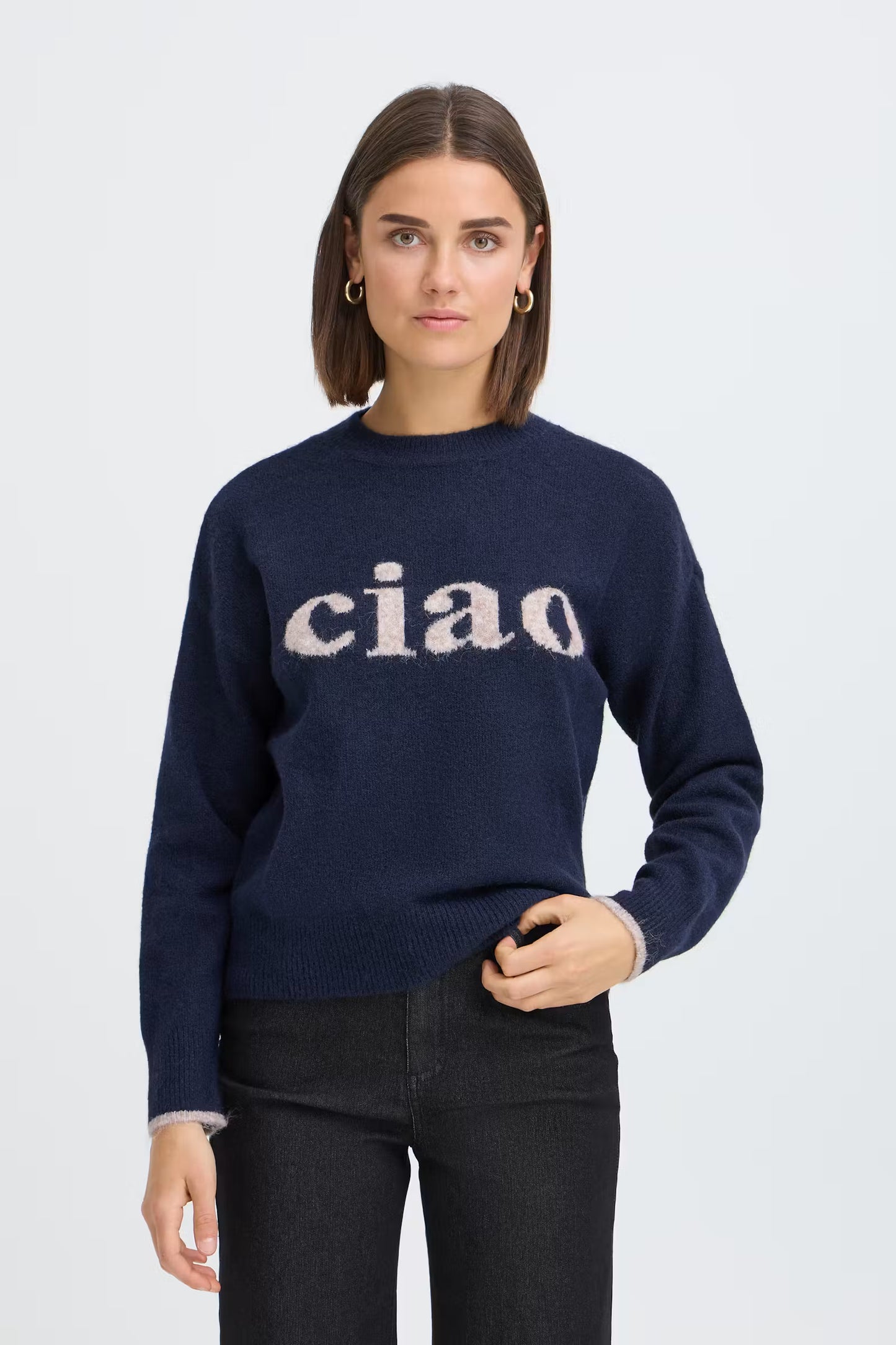 A woman wearing a navy blue knit sweater with the word 'ciao' knitted across the chest in a light beige tone, paired with black jeans, against a light gray studio background.