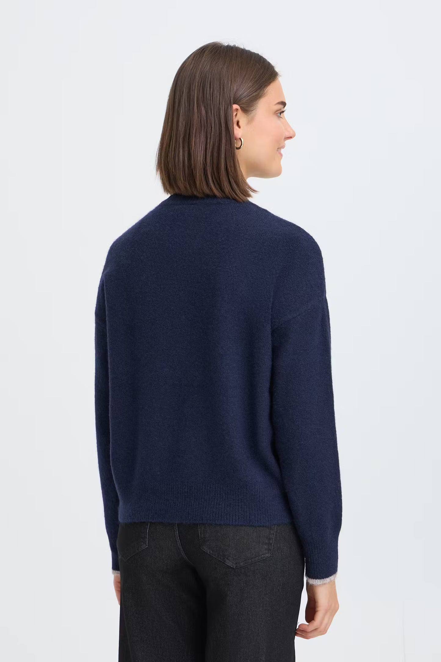 A woman with shoulder-length brown hair is shown from the back wearing a navy blue knit sweater and dark jeans against a light gray studio background.