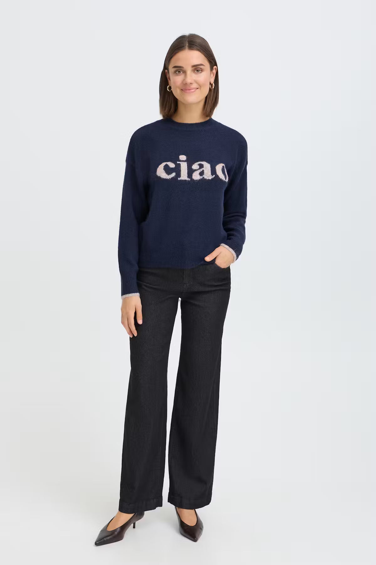 A woman modeling a navy blue knit sweater with 'ciao' written across the chest in white, paired with black wide-leg trousers and dark pointed-toe heels, against a plain light gray studio background.