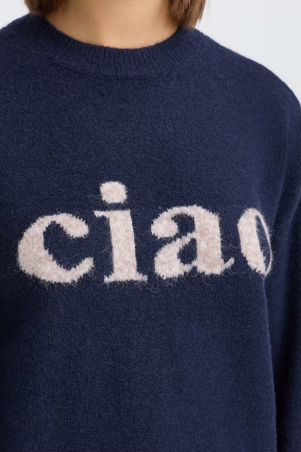 Close-up of a navy blue knit sweater with a fuzzy cream-colored 'ciao' text graphic across the chest, worn by a model.