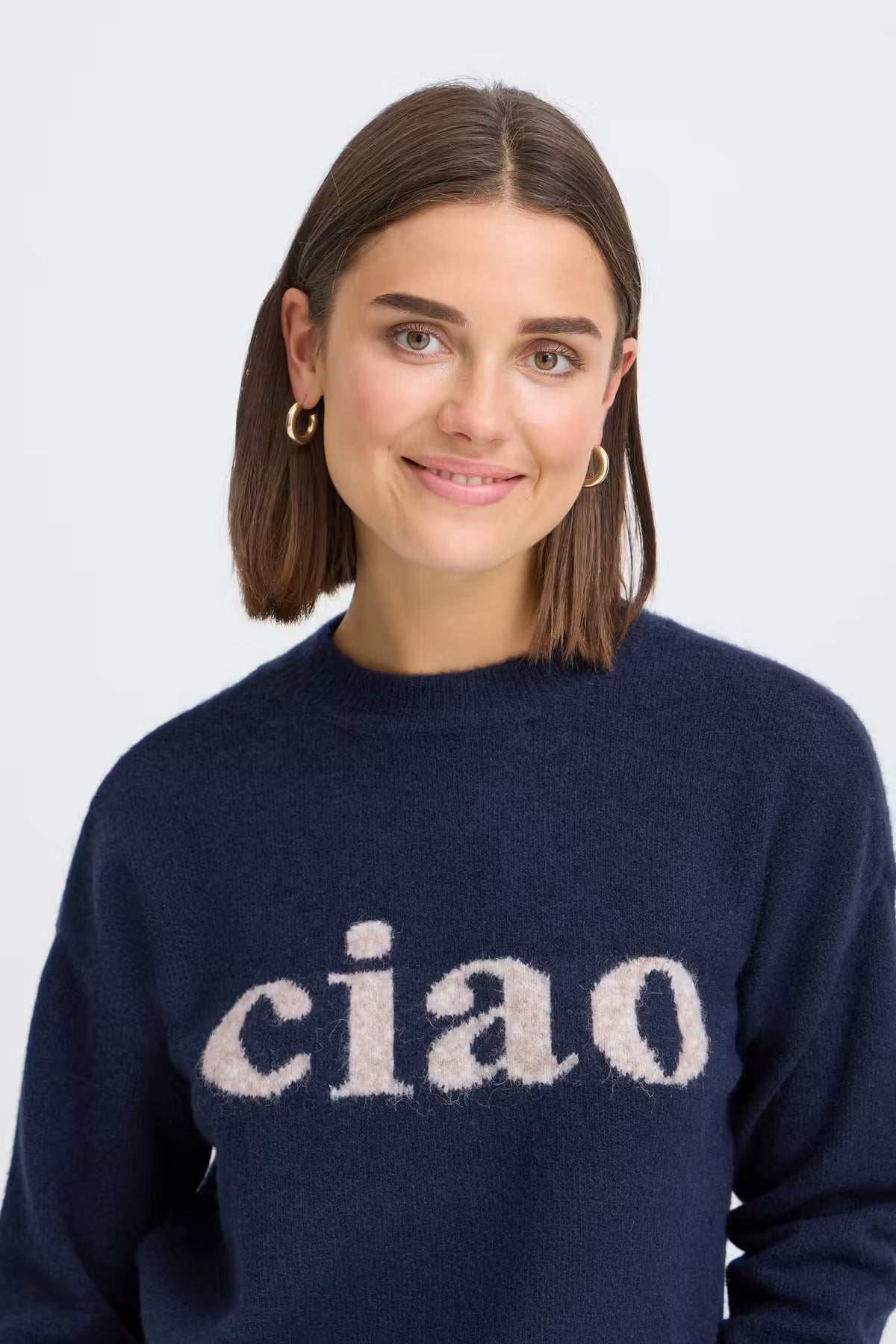 A woman with shoulder-length brown hair wearing a navy blue knit sweater with the word 'ciao' in a fuzzy cream-colored lettering across the chest, set against a plain light gray background.