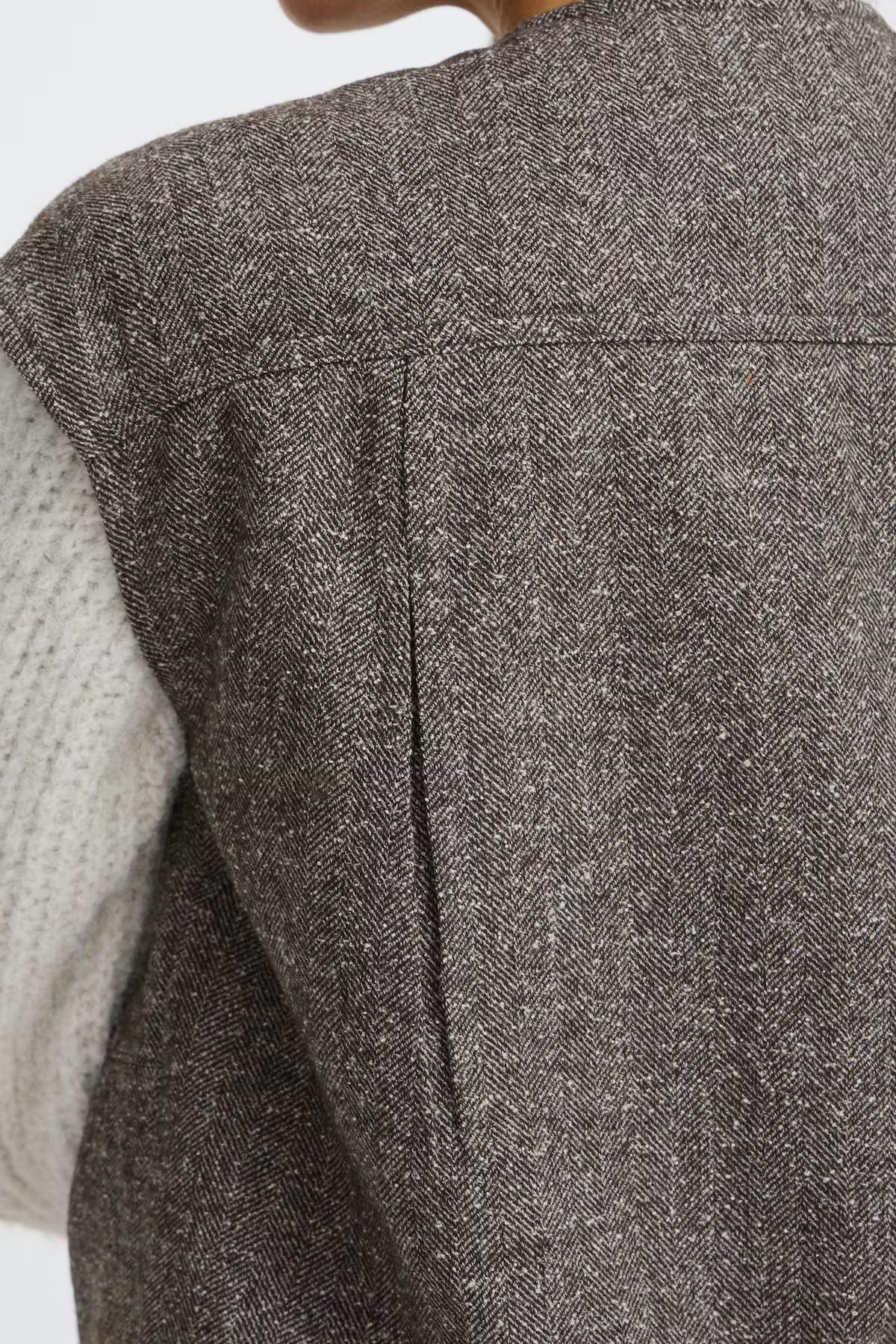 Close-up of a person wearing a dark brown and gray herringbone tweed vest over a light gray knit sweater, showing the back of the vest with a central seam.