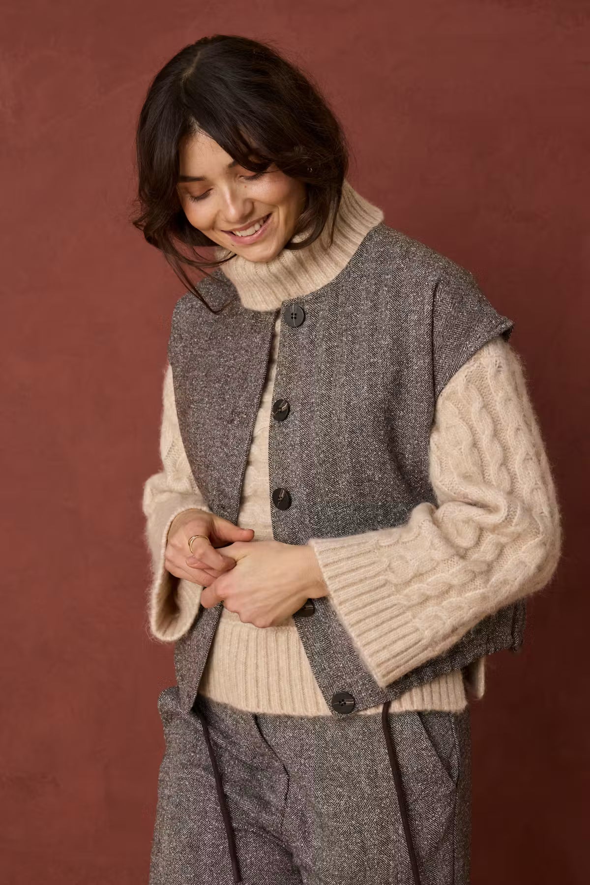 A woman wearing a gray wool-blend vest over a cream cable-knit sweater and matching gray trousers, standing against a warm rust-brown studio background.