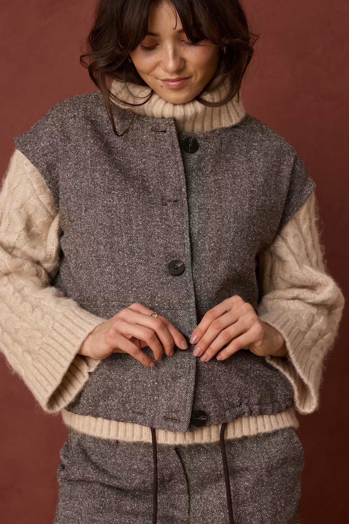 A woman wearing a cropped, sleeveless gray tweed vest with beige knit ribbed trim and matching gray tweed trousers, posed against a solid dark reddish-brown background.