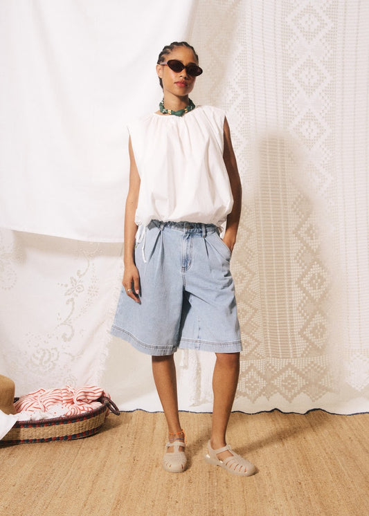 Woman wearing a white sleeveless top and light blue denim shorts against a textured white wall.