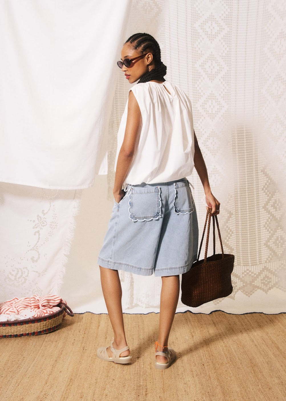 Woman in a white sleeveless top and denim skirt holding a brown bag against a textured white wall.