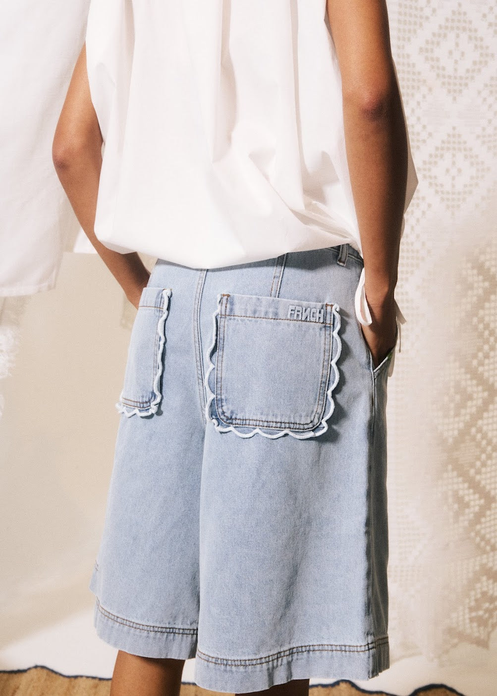 Person wearing light blue denim shorts with scalloped pocket details on a neutral background