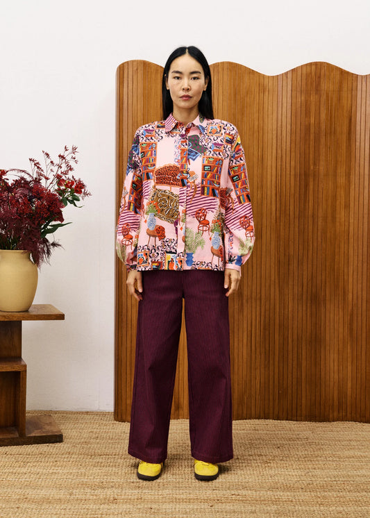 Person wearing a colorful patterned shirt and maroon pants standing in front of a wooden screen.