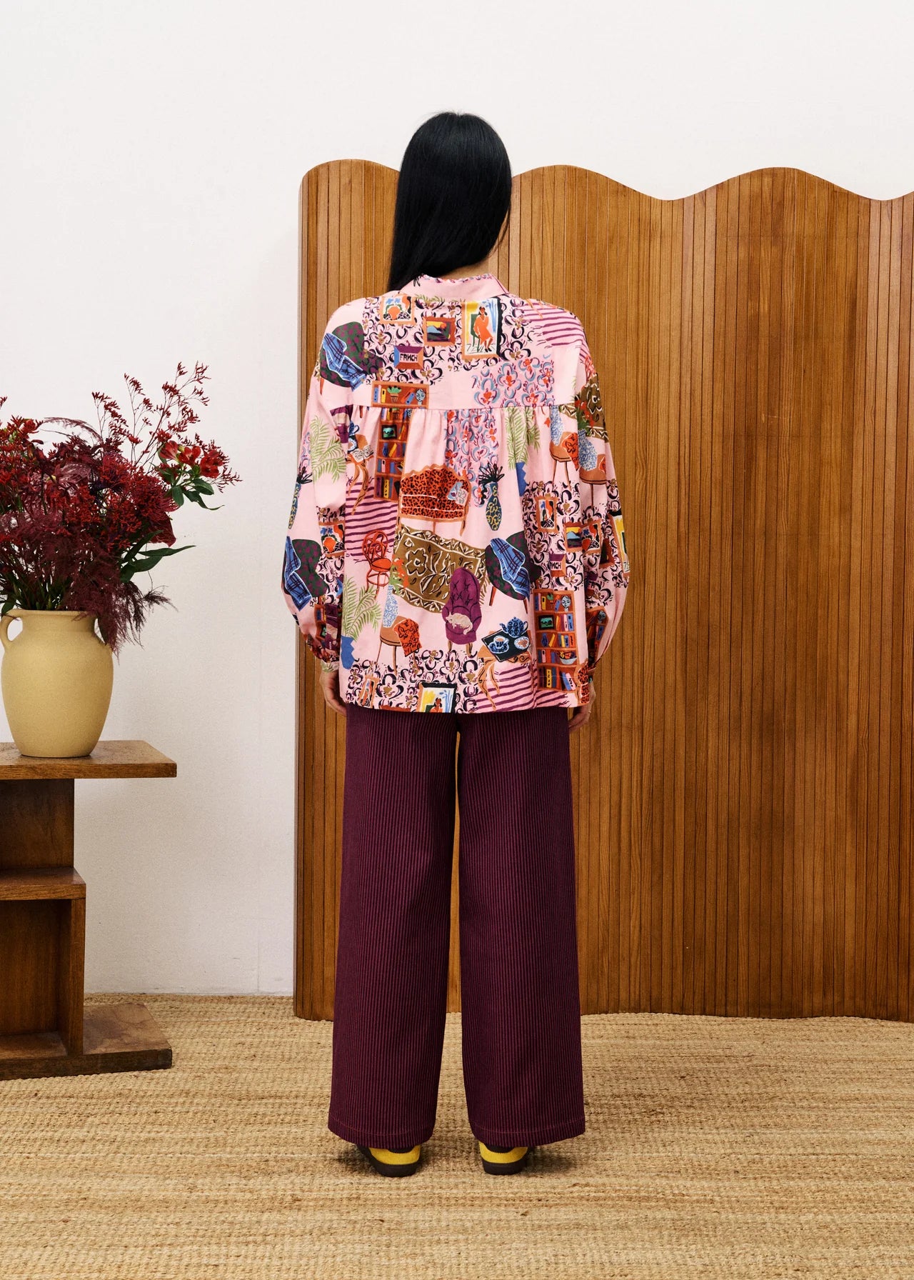 Person wearing a colorful patterned jacket and purple pants standing in front of a wooden screen.