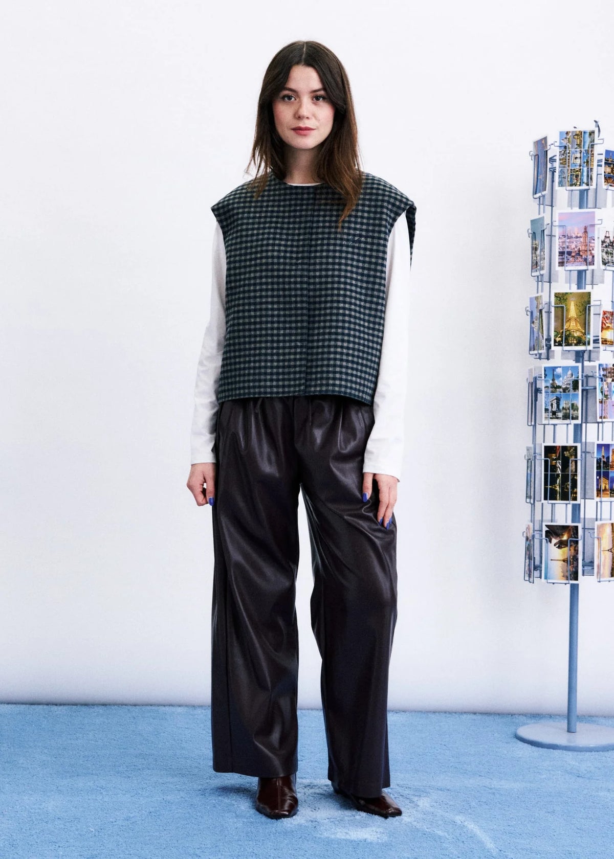 A model stands in a studio wearing a sleeveless black and white houndstooth vest over a white long-sleeve shirt, paired with black wide-leg satin pants and dark brown shoes. A white wall and a blue floor are visible, with a magazine rack on the right.