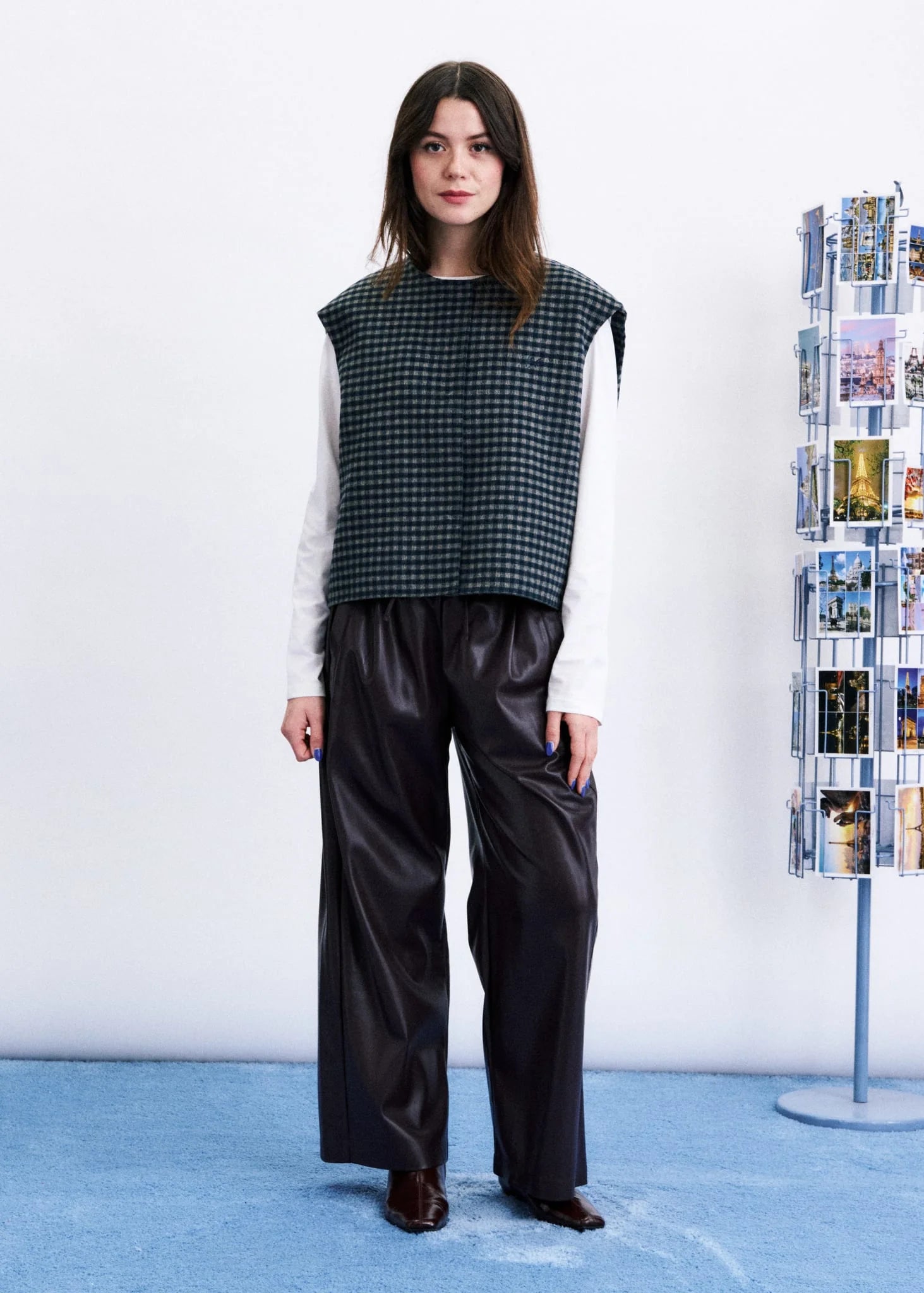 A model stands in a studio wearing a sleeveless black and white houndstooth vest over a white long-sleeve shirt, paired with black wide-leg satin pants and dark brown shoes. A white wall and a blue floor are visible, with a magazine rack on the right.