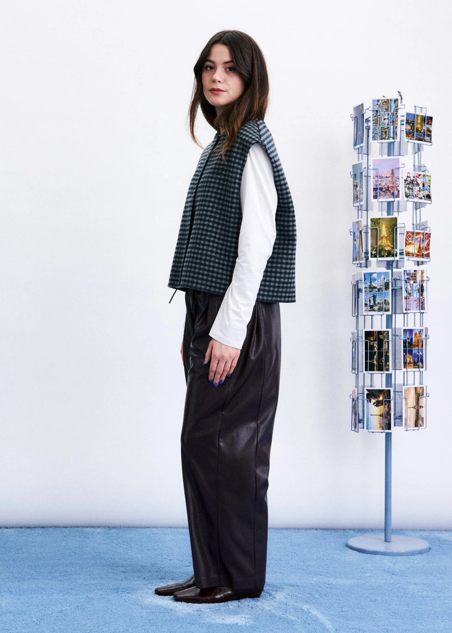 A model stands in profile wearing a sleeveless black and green houndstooth vest over a white long-sleeve shirt and dark wide-leg trousers. She is standing in a studio with a light blue floor against a white wall, next to a tall display stand filled with small printed photos.