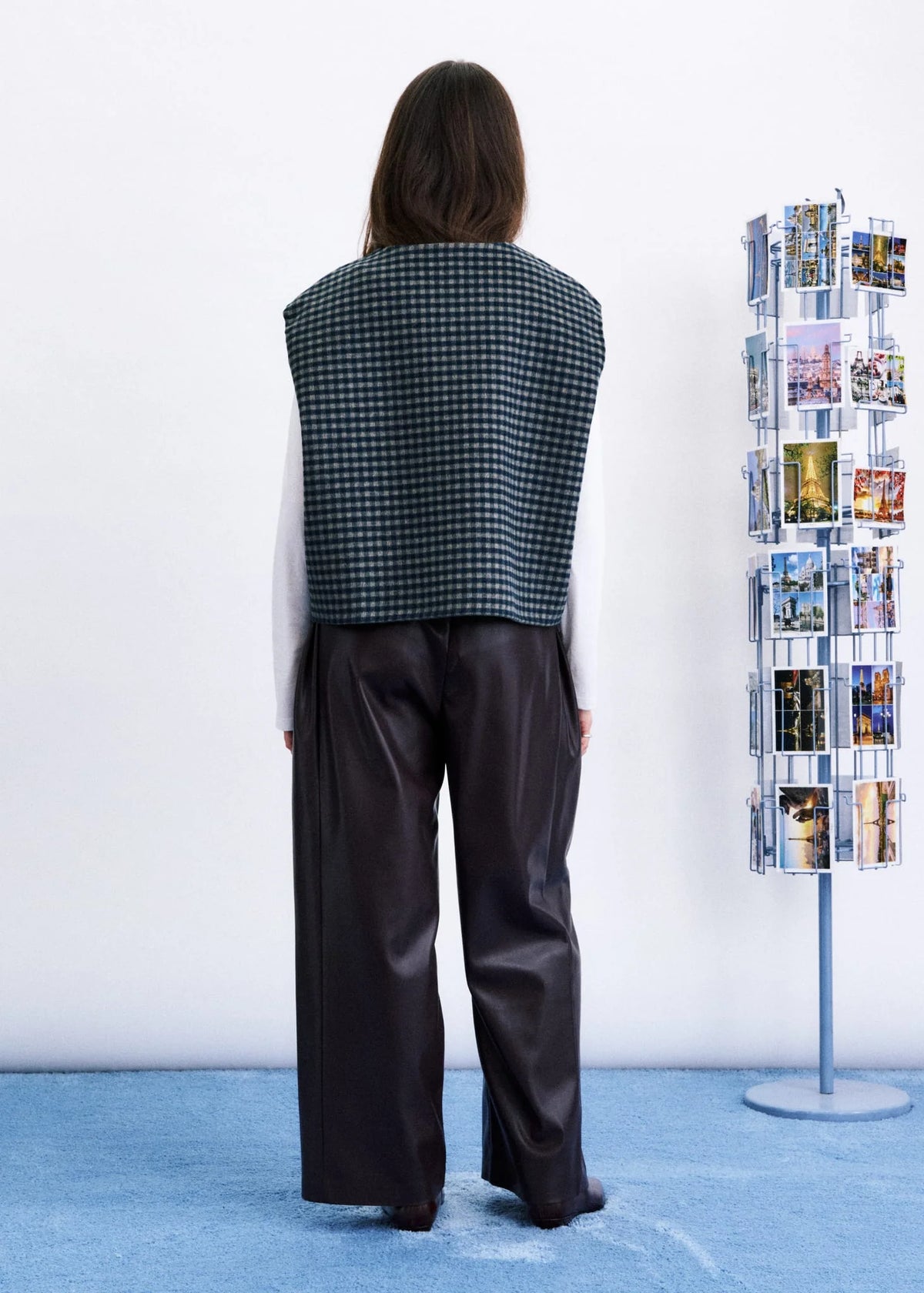 A model viewed from behind wearing a sleeveless black and white gingham vest over a white long-sleeve top and dark wide-leg trousers, standing in a studio with a blue floor against a white wall. A clothing rack with photos is visible to the right.