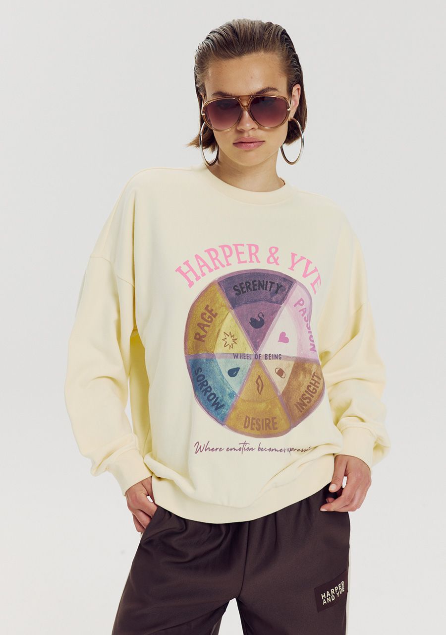 A model wearing a cream-colored long-sleeve sweatshirt with a colorful graphic print on the front that reads 'HARPER & YVES' and 'Serenity Peace Love Beauty'. The sweatshirt is paired with dark brown sweatpants and sunglasses, set against a plain light gray background.