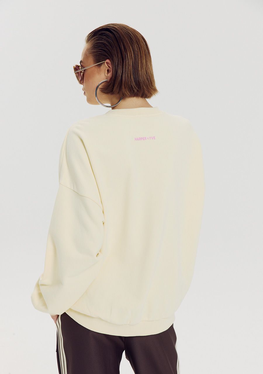 A model from the back wearing a cream-colored crewneck sweatshirt with a small pink 'SWEATERS-OUTLET' logo on the upper back, paired with dark brown track pants with white side stripes, against a light gray background.