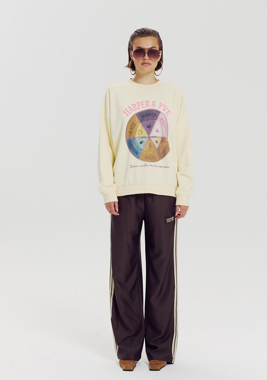 A model stands against a light gray studio background wearing a cream sweatshirt with a colorful circular graphic and the words 'HAMMER & LOVE' across the chest, paired with dark brown track pants featuring white side stripes and brown shoes.