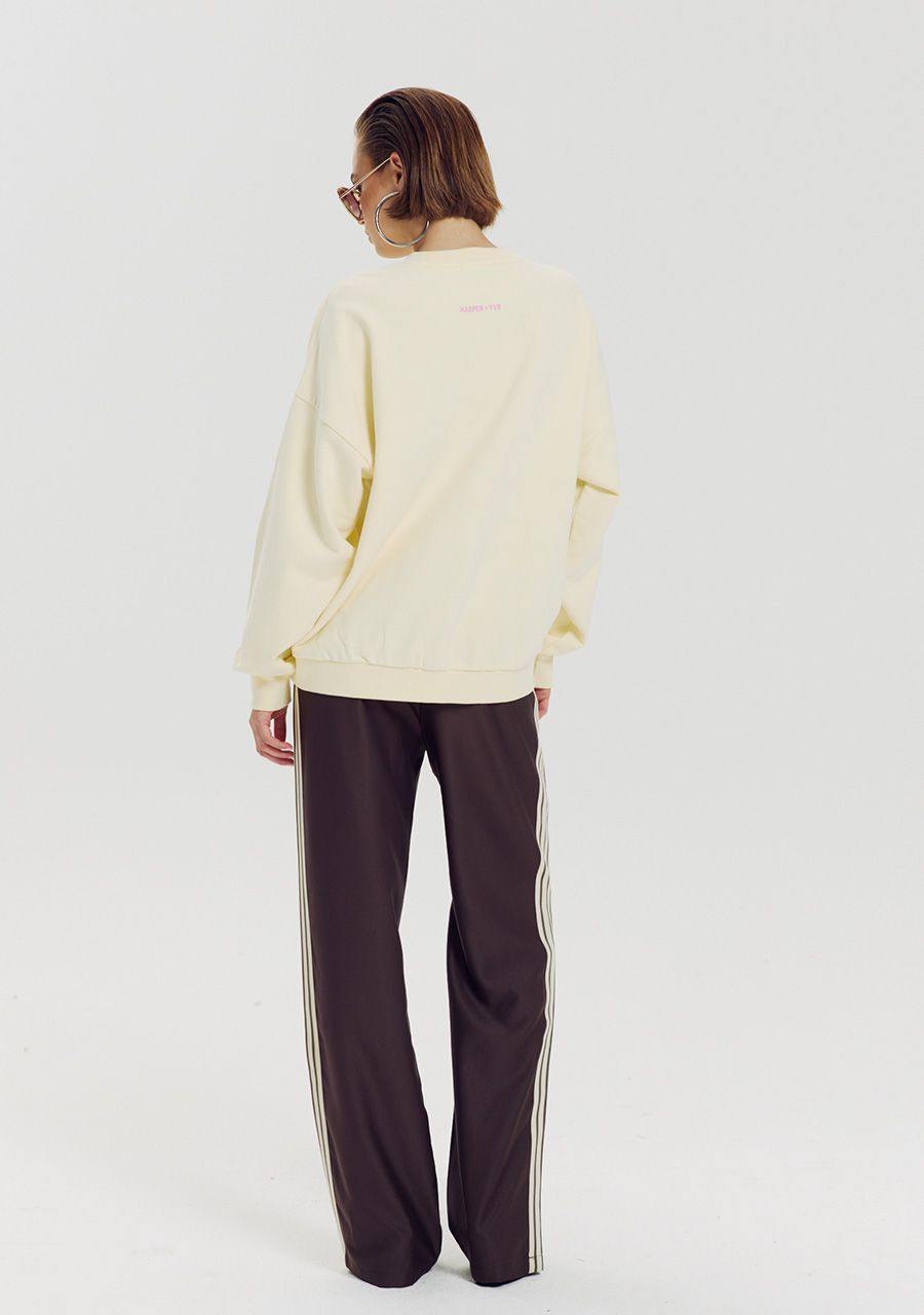 A model viewed from the back wearing a cream-colored crewneck sweatshirt and dark brown track pants with white side stripes, set against a plain light gray studio background.