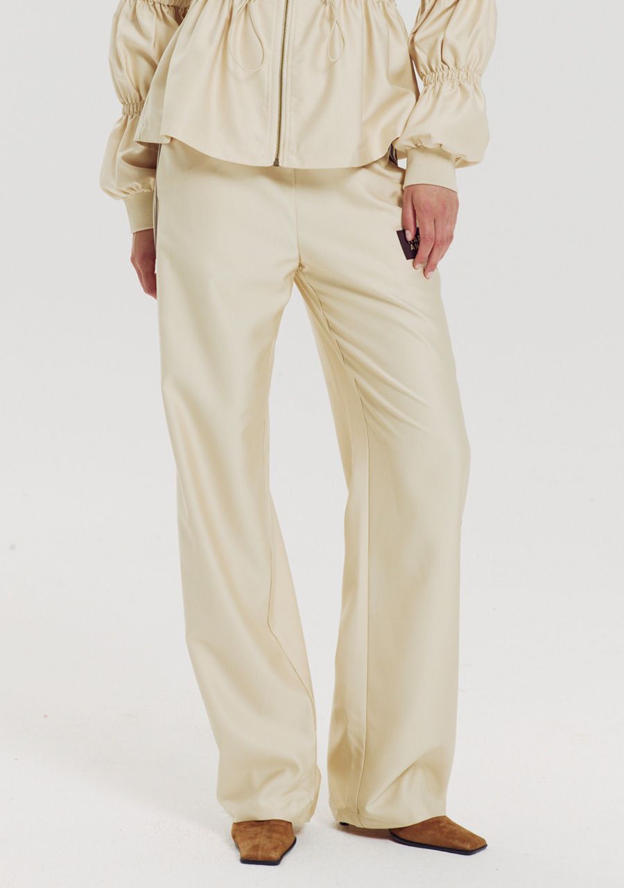 A model wearing a matching cream-colored two-piece outfit consisting of a cropped long-sleeve top with gathered cuffs and wide-leg trousers, paired with brown pointed-toe flats, against a plain white studio background.