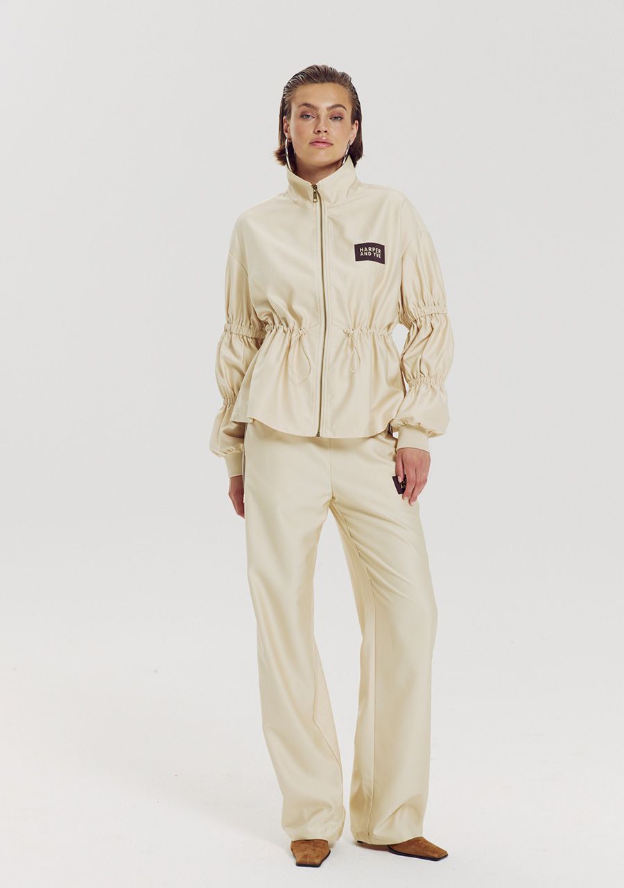 A model stands against a plain light gray studio background wearing a matching cream-colored two-piece tracksuit with a zip-up jacket featuring gathered sleeves and a small black rectangular patch on the chest, paired with matching wide-leg pants.