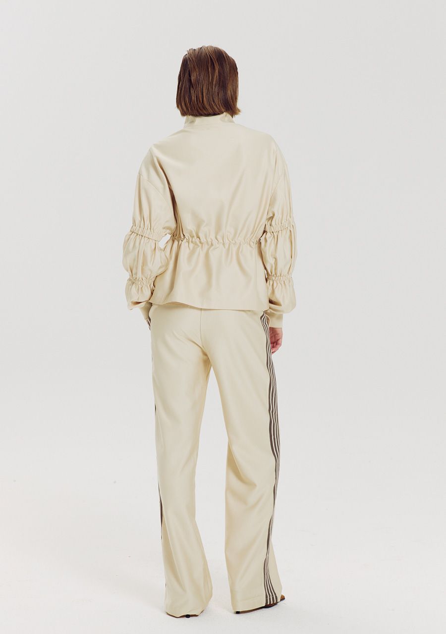 A model viewed from the back wearing a matching beige two-piece tracksuit with a cropped, gathered long-sleeve top and wide-leg pants featuring dark side stripes.