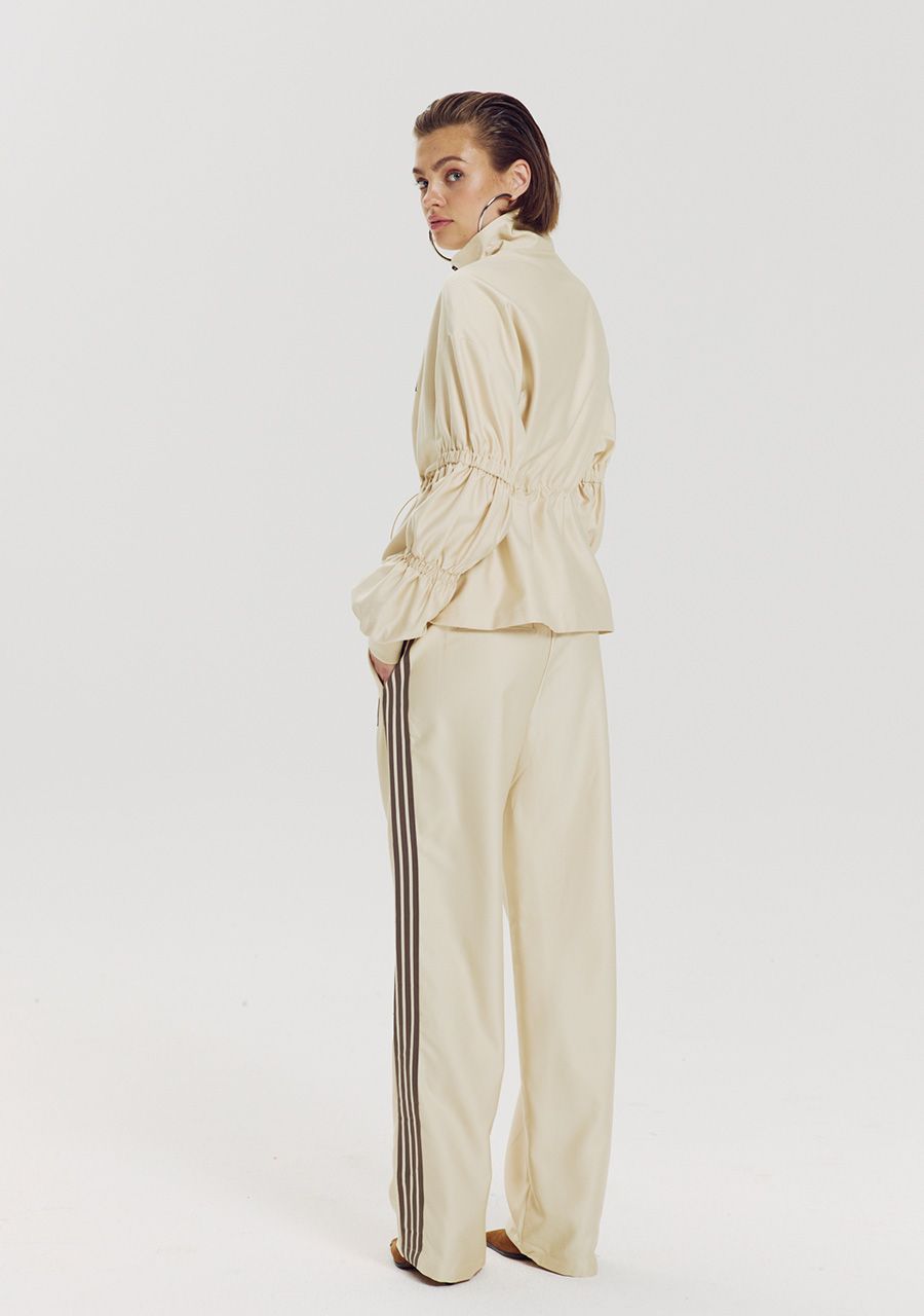 A model is shown from a side-back angle wearing a cream-colored two-piece tracksuit with a belted jacket and wide-leg pants featuring dark brown stripes down the side, set against a plain light gray studio background.