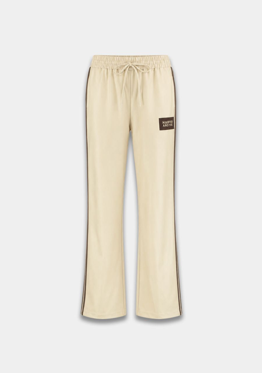 Cream-colored wide-leg sweatpants with dark brown side stripes and a small dark brown 'PASTRY CRAFTS' brand patch on the left thigh, shown against a light gray background.