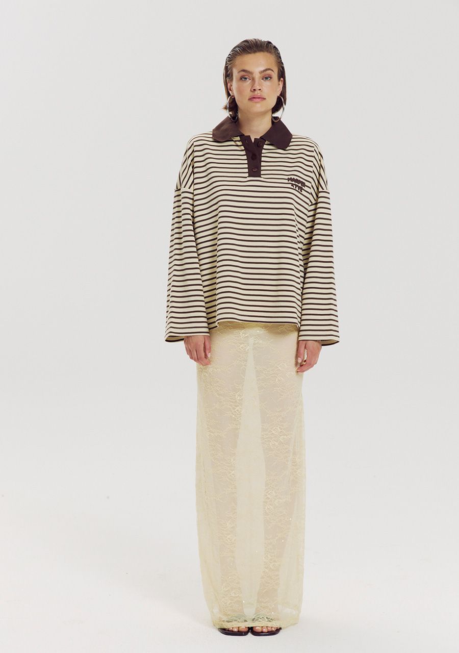 A model stands against a plain light gray studio background wearing a long-sleeve polo shirt with horizontal dark brown and beige stripes, a dark brown collar, and a small chest logo, paired with wide-leg cream trousers.