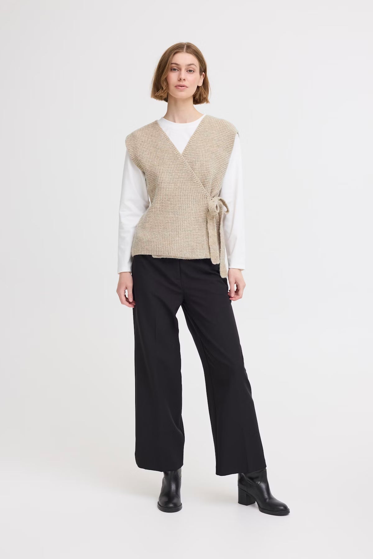 Woman wearing a beige vest over a white shirt with black pants on a white background