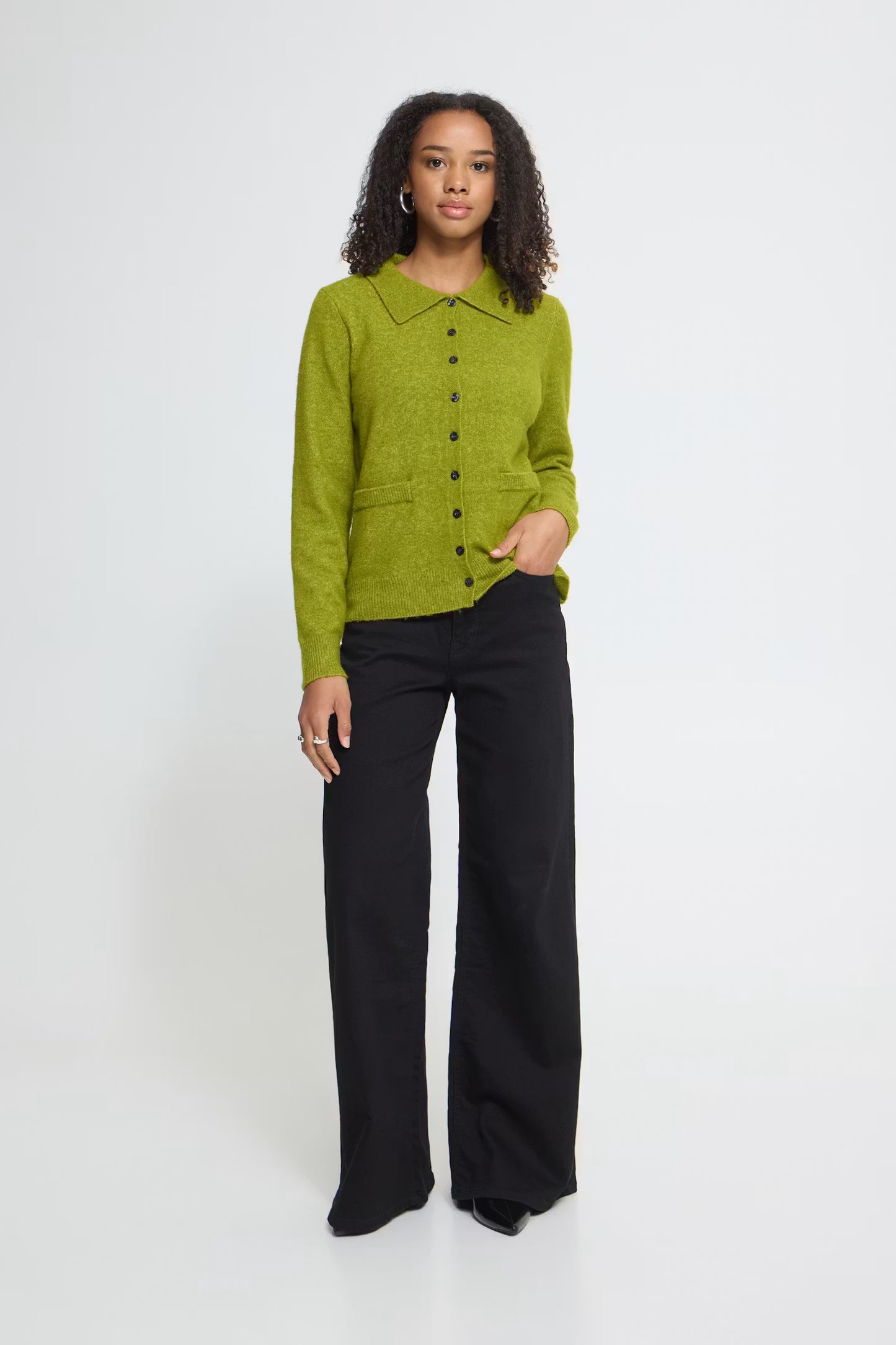 A woman stands against a plain light gray studio background wearing a bright olive green button-front knit cardigan with a collar over black wide-leg trousers.
