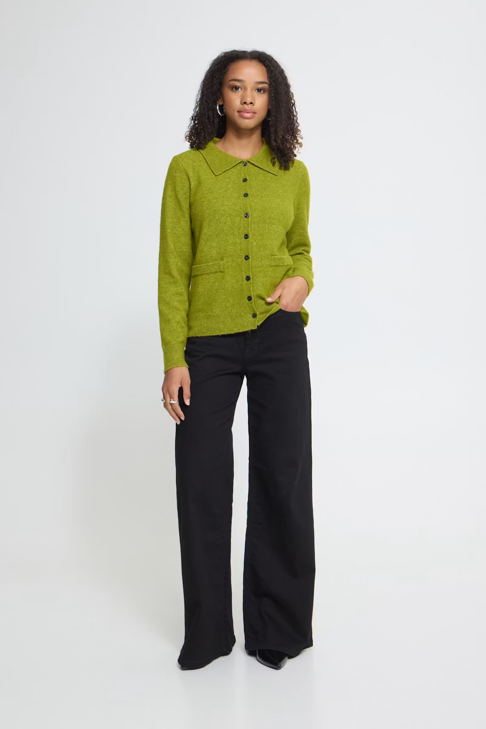A woman stands against a plain light gray studio background wearing a bright olive green button-front knit cardigan with a collar over black wide-leg trousers.