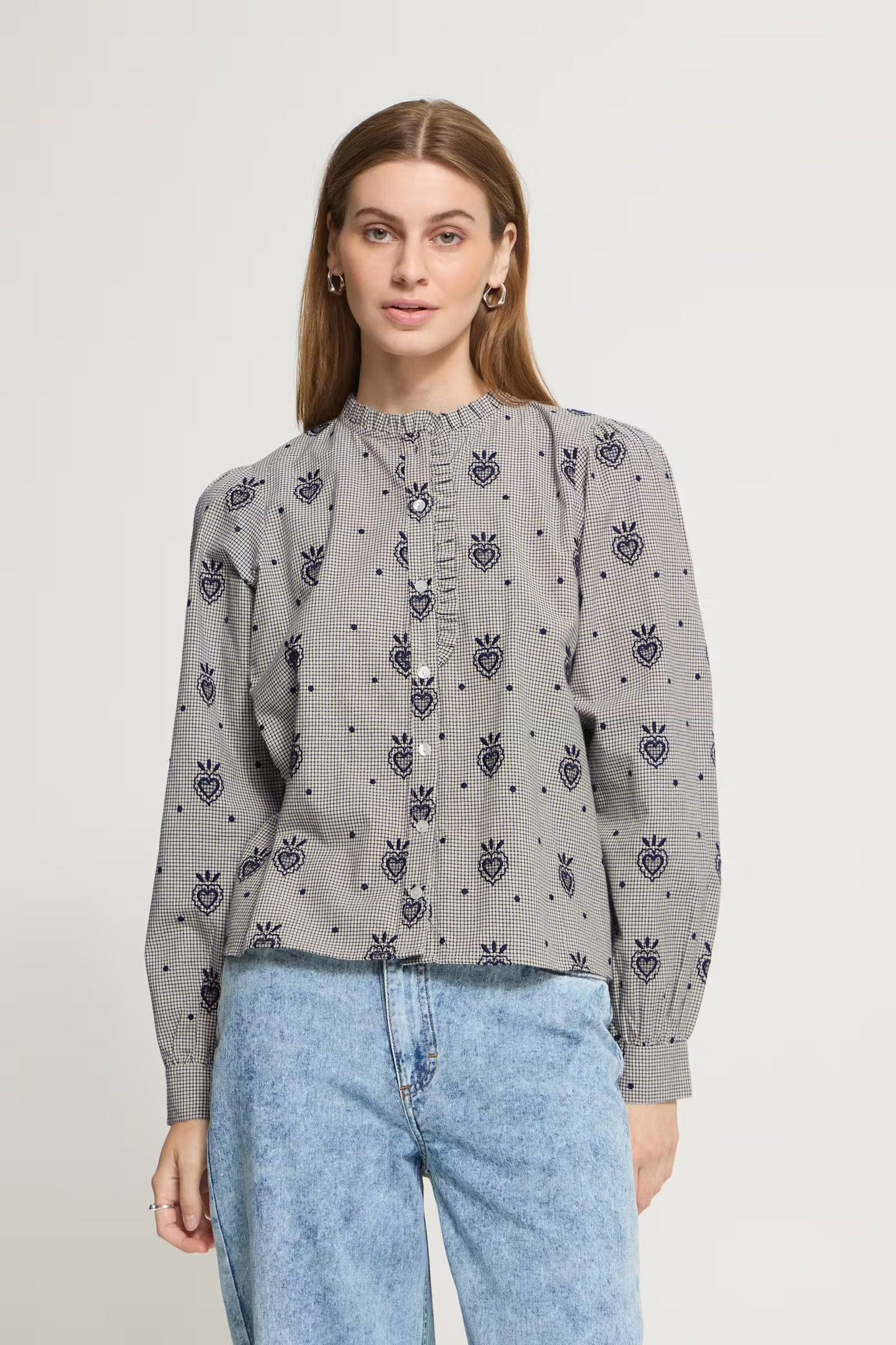 A woman wearing a gray long-sleeve blouse with a repeating dark blue embroidered heart and crown motif, paired with light blue jeans, against a plain off-white background.
