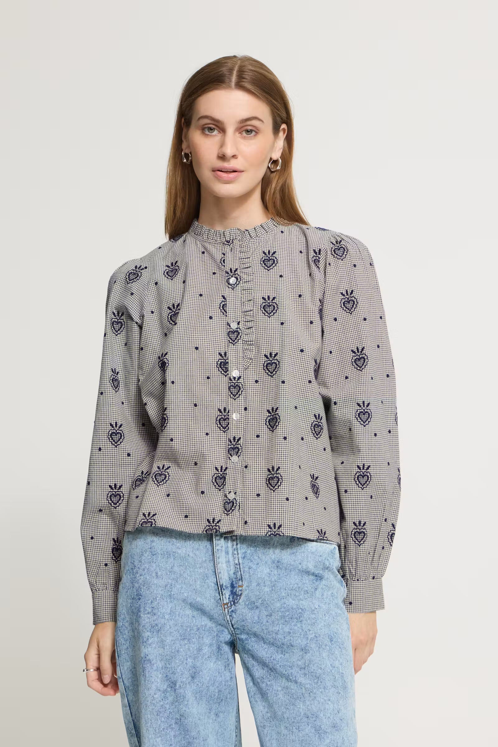 A woman wearing a gray long-sleeve blouse with a repeating dark blue embroidered heart and crown motif, paired with light blue jeans, against a plain off-white background.