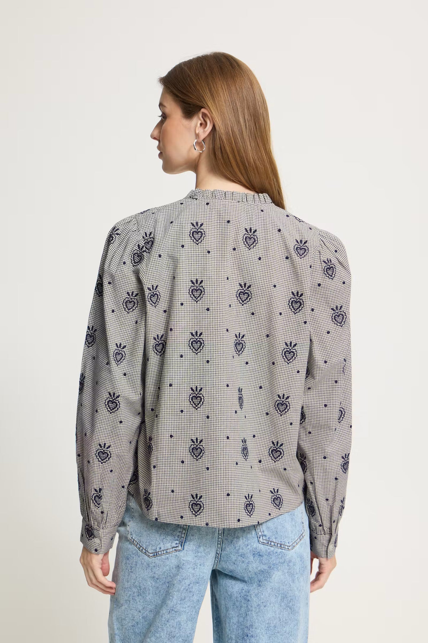 A woman shown from the back wearing a gray long-sleeve blouse with a repeating dark blue embroidered pineapple and crown motif, paired with light blue jeans, against a plain off-white background.