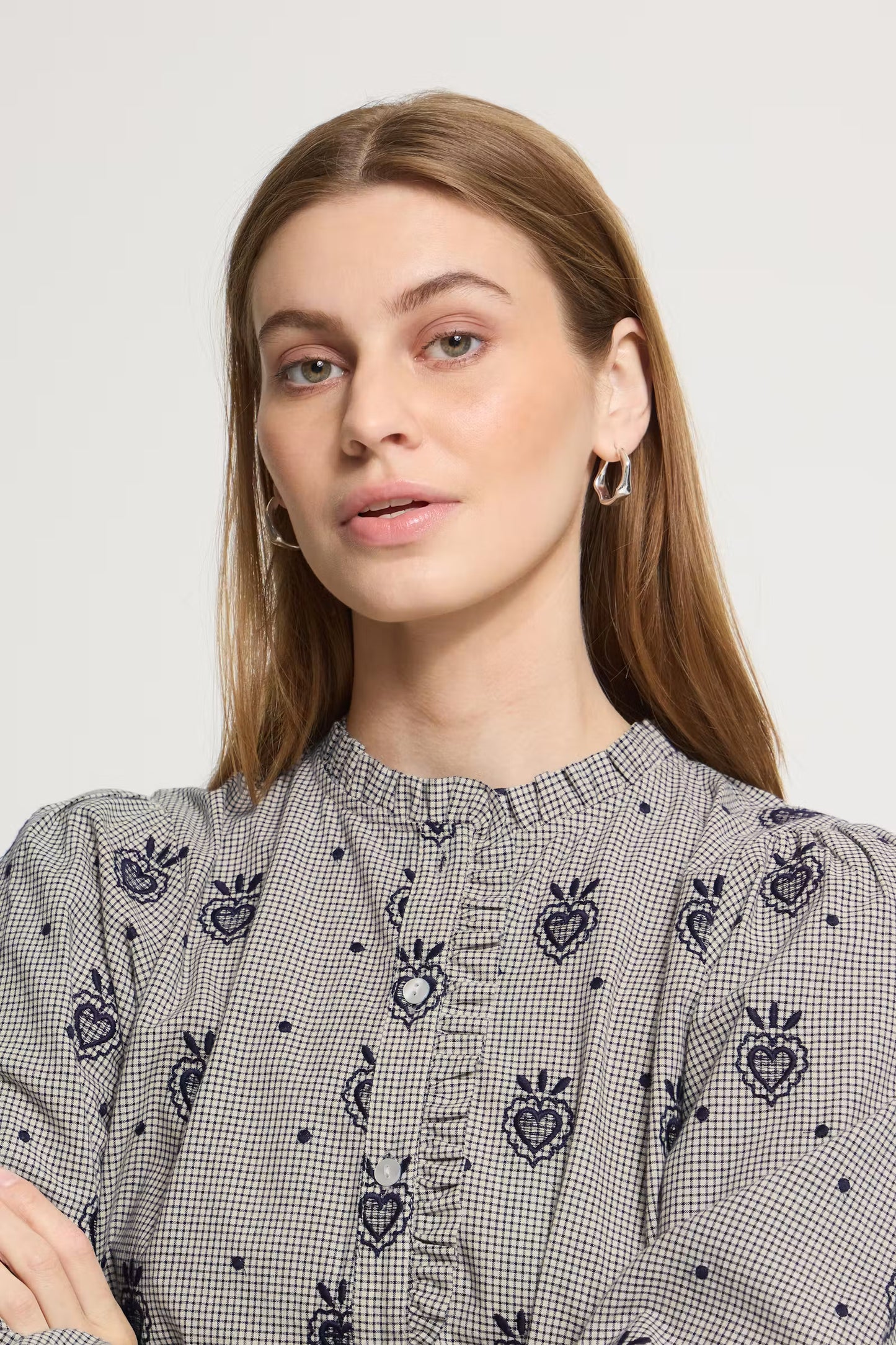 A woman wearing a gray and white gingham blouse with dark blue embroidered heart motifs and a ruffled high neckline, shown against a plain light gray background.