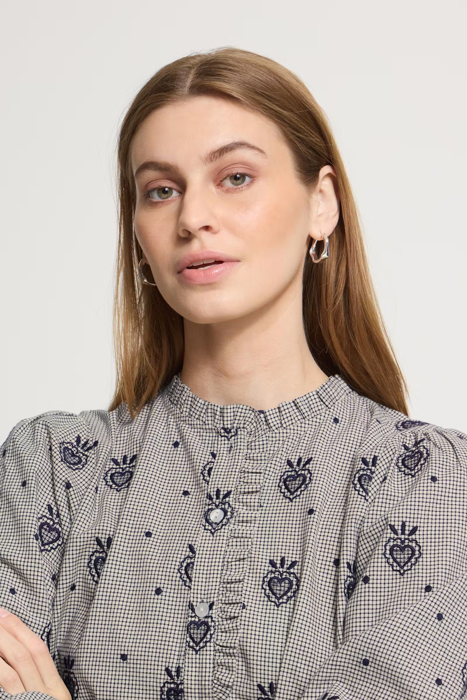 A woman wearing a gray and white gingham blouse with dark blue embroidered heart motifs and a ruffled high neckline, shown against a plain light gray background.