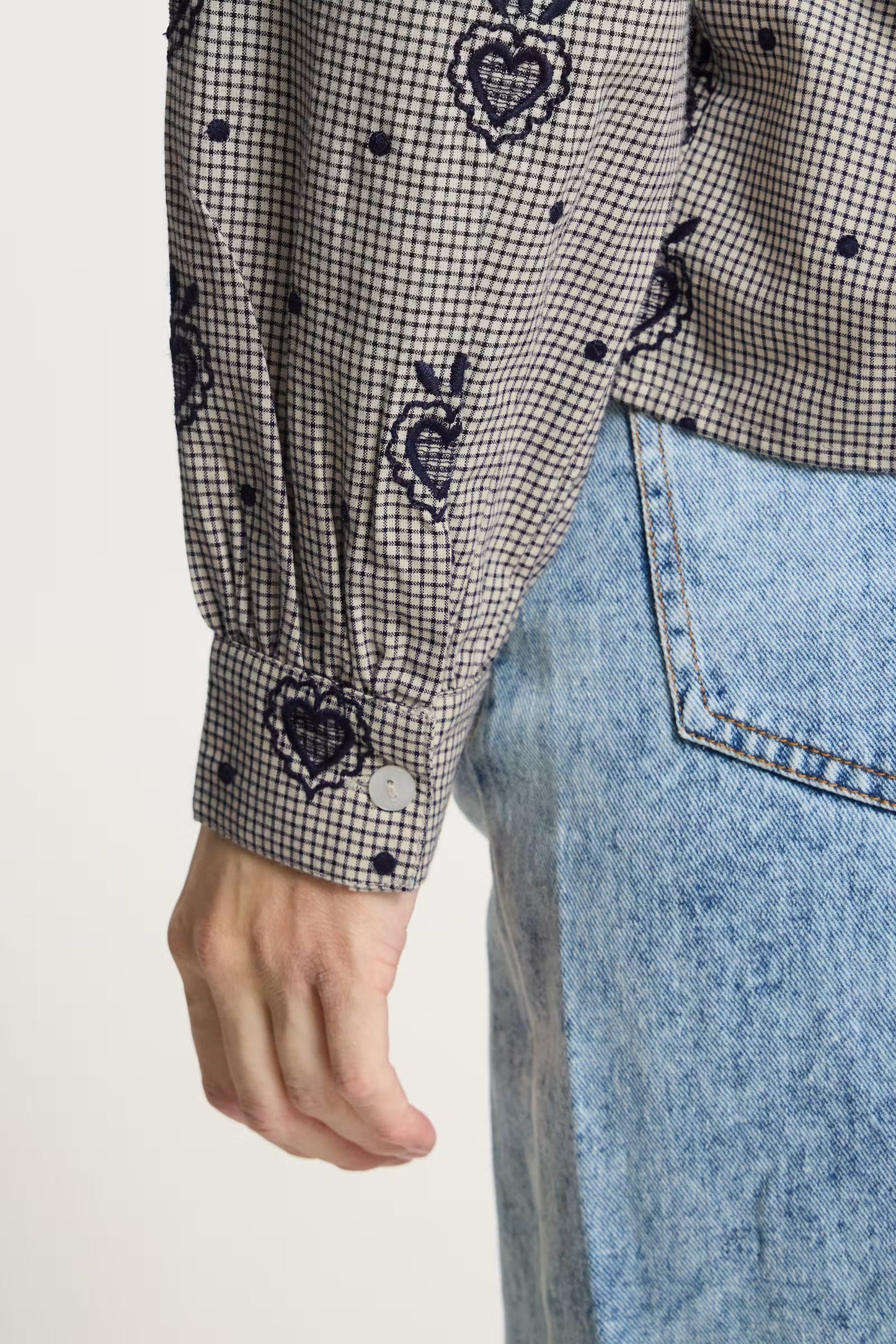 Close-up of a person wearing a black and white gingham long-sleeve shirt with embroidered heart motifs and light blue denim jeans against a plain light background.
